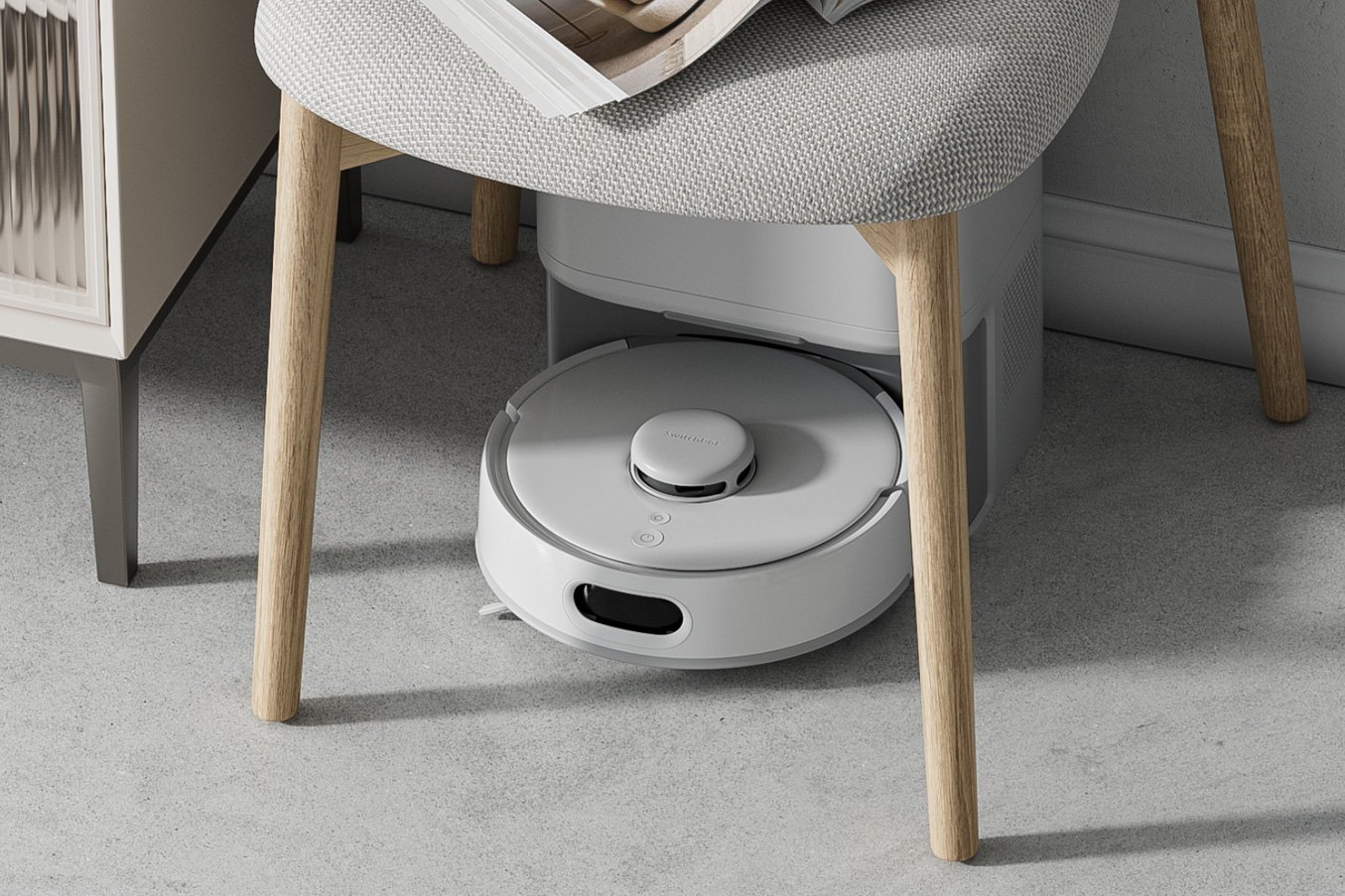 A SwitchBot Mini Robot Vacuum K10+ Pro docked at its base station under a chair.