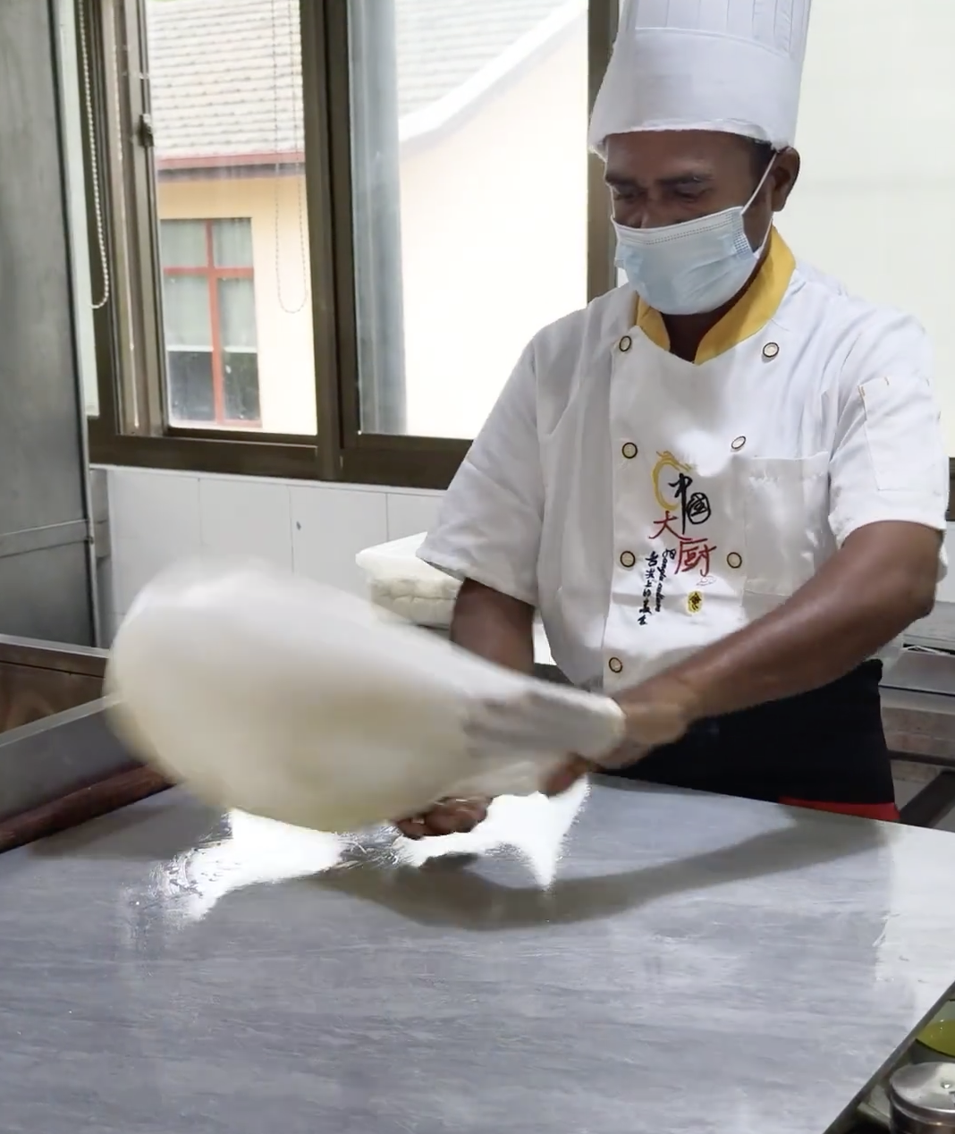 Chef wearing a mask and white uniform stretching a large sheet of dough on a work surface in a bright kitchen, illustrating traditional Chinese cooking techniques.